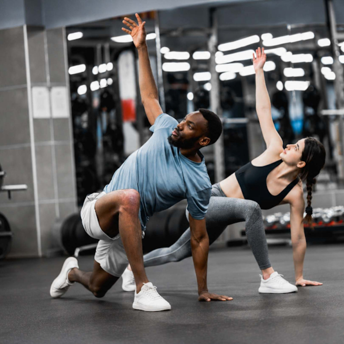 A man and a woman in athletic wear perform a synchronized yoga or stretching pose in a gym. The man wears a light blue t-shirt, white shorts, and white sneakers, while the woman wears a black sports bra, gray leggings, and white sneakers.