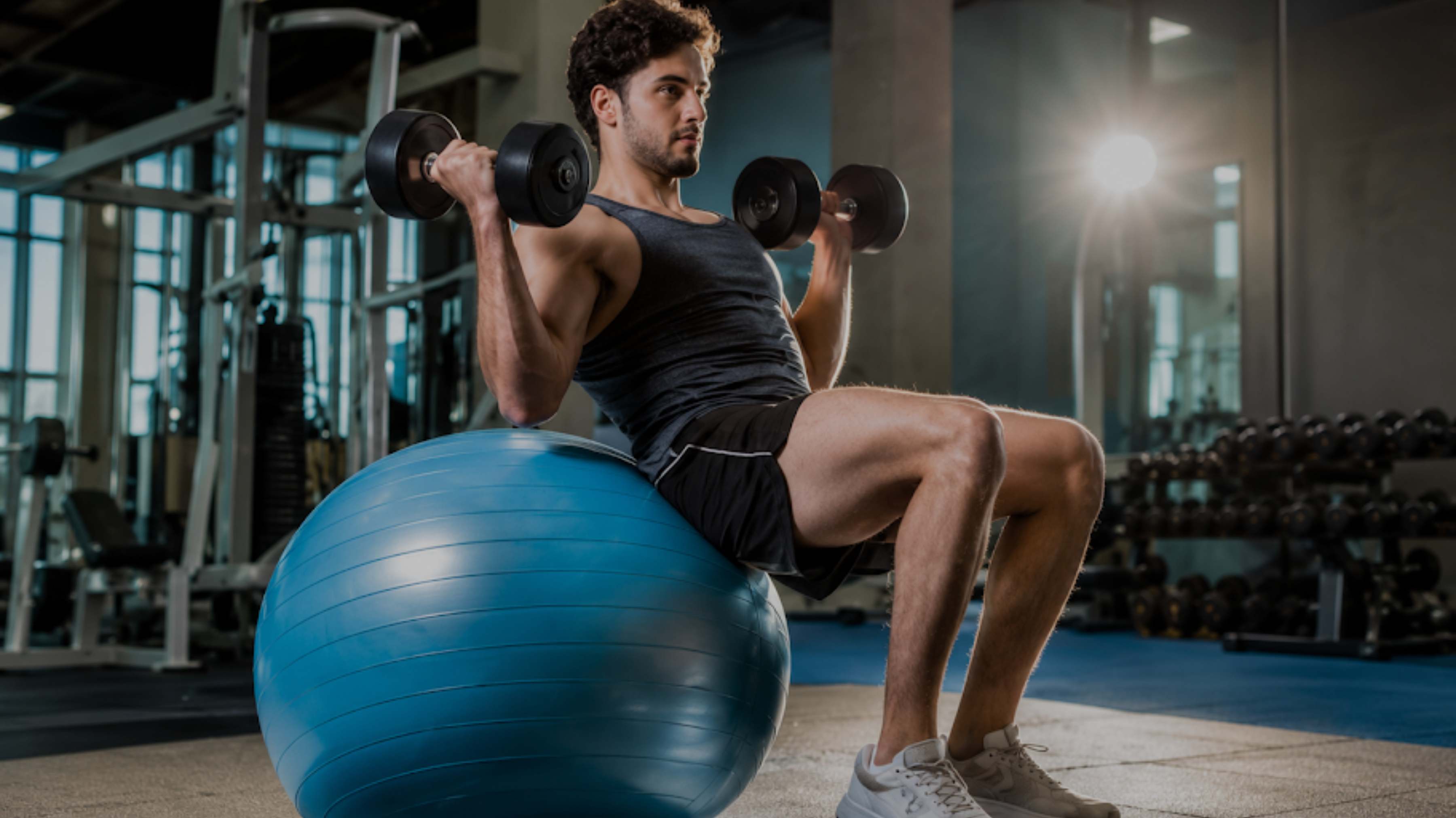 A man in a gym sits on a blue exercise ball while lifting two black dumbbells overhead. He is wearing a black tank top, black shorts, and white sneakers.