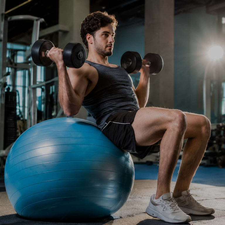A man in a gym sits on a blue exercise ball while lifting two black dumbbells overhead. He is wearing a black tank top, black shorts, and white sneakers.