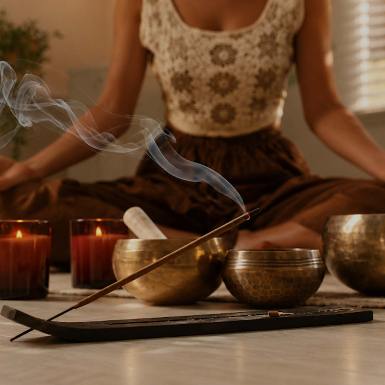 A person in a patterned top and brown skirt sits cross-legged in a meditative pose with hands in prayer position, surrounded by brass singing bowls, lit candles, and a smoking incense stick in a warm, softly lit indoor setting.