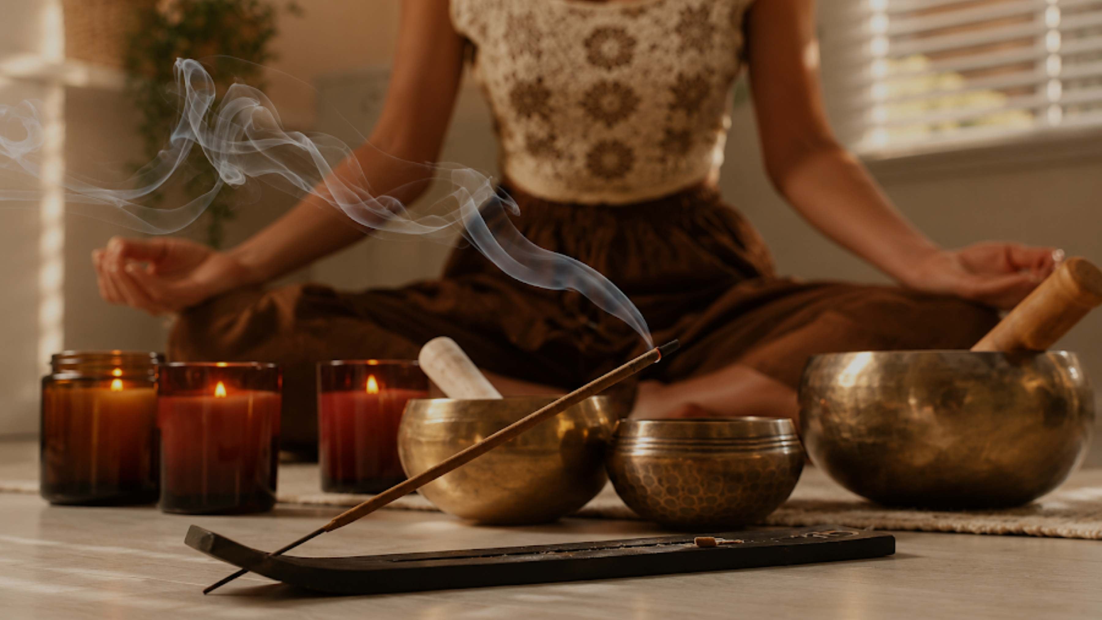 A person in a patterned top and brown skirt sits cross-legged in a meditative pose with hands in prayer position, surrounded by brass singing bowls, lit candles, and a smoking incense stick in a warm, softly lit indoor setting.