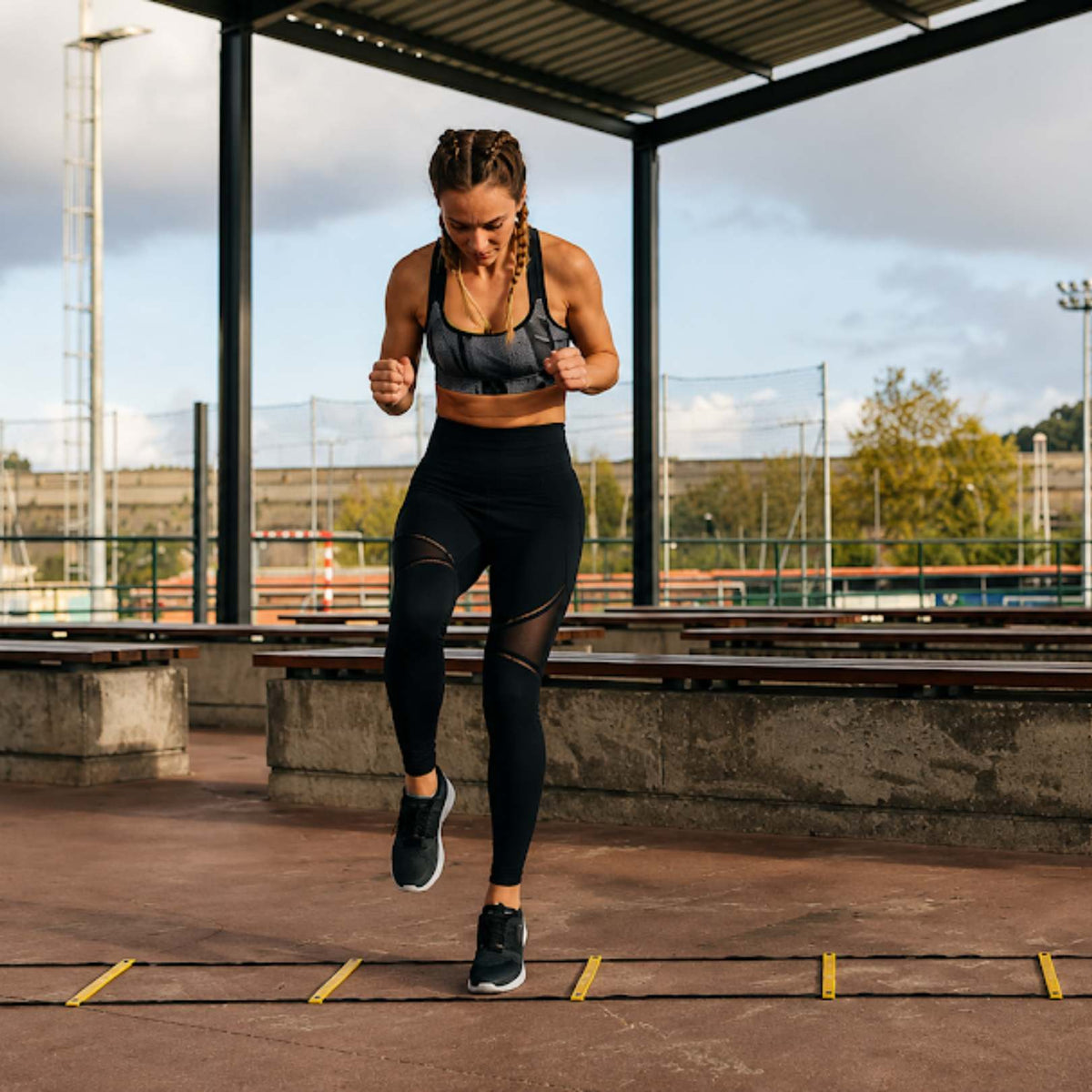 A woman in athletic wear, including a gray and black sports bra and black leggings, is running agility drills on a yellow ladder on an outdoor track under a covered structure.